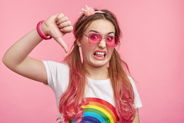 Fashionable woman shows disapproval sign, gives thumb down gesture, dislikes something, has disgusting expression, isolated over pink background. Negative human expression and body language concept.