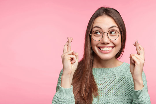 Indoor shot of glad brunette woman wears spectacles, smiles broadly, keeps fingers crossed, hope for good luck during long trip, isolated over pink background with copy space for your text or hearder
