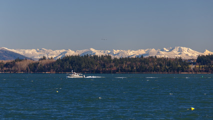 Fototapeta premium A winter view across Padilla Bay with boat passing by, evergreen trees by the shoreline, and snow covered mountains in the background.