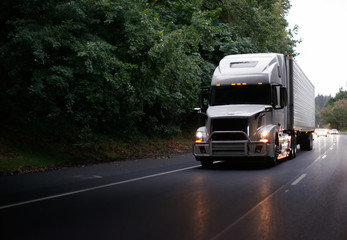 Fototapeta na wymiar Modern big rig semi truck with guard and turn on headlights and reefer semi trailer on evening road