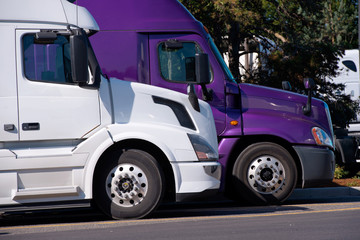 White and purple big rigs semi trucks stand on truck stop side by side