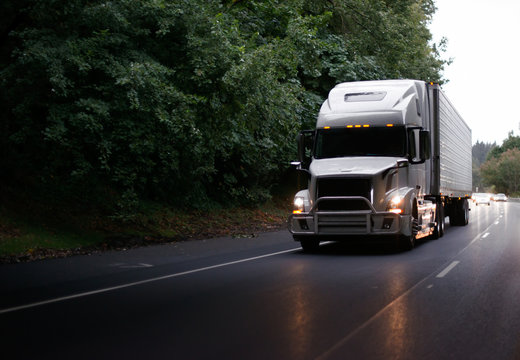 Modern big rig semi truck with guard and turn on headlights and reefer semi trailer on evening road
