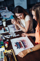 Fototapeta premium Two young women attending aquarelle painting classes for adults at art school. A girl showing her artwork to friend.