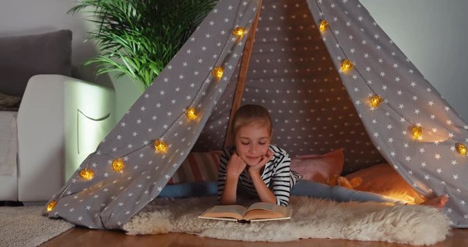 Cheerful child girl 9 years old reading book in wigwam. Dolly shot