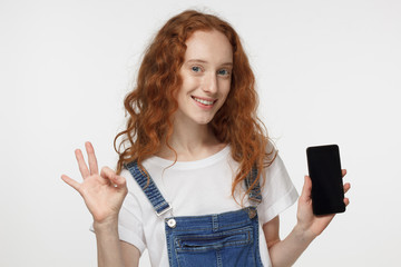 Closeup shot of young attractive redhead European woman isolated on white background showing blank cellphone screen and OK sign with fingers as if approving service or product, copyspace provided