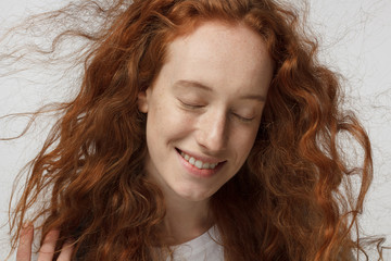 Detailed indoor headshot of young attractive teenage girl isolated on white background, her red hair flowing in air in waves, her eyes closed with pleasure, smiling gently in relaxed atmosphere