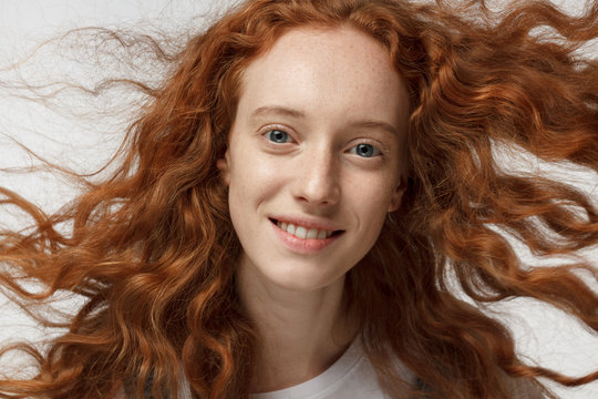 Detailed horizontal photo of young beautiful redhead European girl isolated on white background, her curly hair waving in torrent of air as she is posing with happy smile, looking pure and feminine