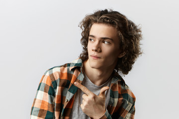 Check this out. Close up portrait of cool fashionable young man with curly hair pointing index finger sideways, indicating copy space on blank studio wall for your text or advertising content