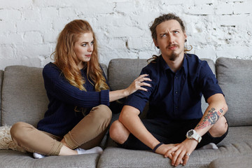 Stylish young couple with facial piercing having disagreement in living room. Unshaven man with tattoo and dreadlocks rolling eyes, feeling irritated with his annoying wife who is asking for money - obrazy, fototapety, plakaty