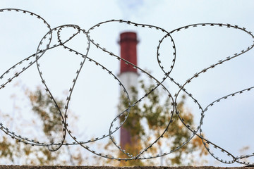 A pipe with smoke behind a fence with barbed wire.