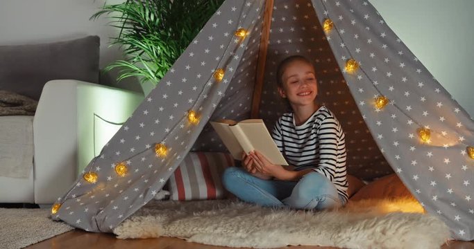 Cheerful child reading book in wigwam and smiling at camera