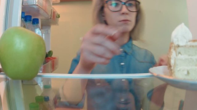 Young woman opens the fridge takes fresch apple and then notices a cake and takes it