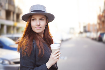 Close up portrait of Young stylish beautiful woman in hat with coffee in hand while walking in the big city streets.