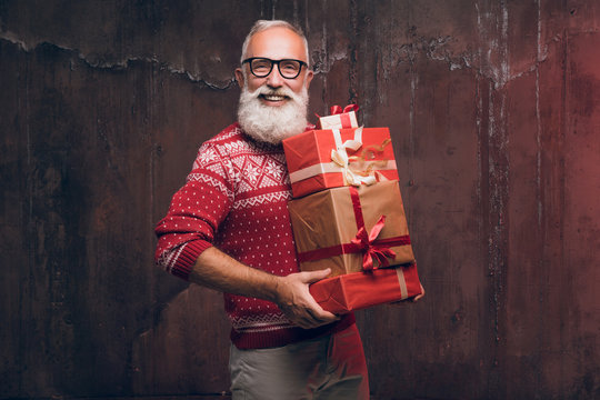 Handsome senior bearded man holding gift box over dark background and looking at camera. Santa Claus wishes Merry Christmas and a happy new year.