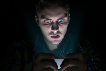 The guy in the shirt sits in the dark and looks at the mobile phone. His face has emotions of surprise