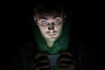 The guy in the shirt sits in the dark and looks at the mobile phone. His face has emotions of surprise