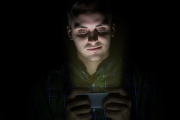 The guy in the shirt sits in the dark and looks at the mobile phone. On the face is the emotion of...