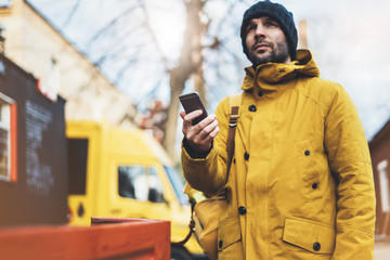 Hipster with yellow backpack, jacket, cap, coffee of thermo cup holding in the hands smartphone, freelance using gadget mobile in autumn street outdoor, tourist man typing message, online process