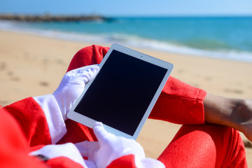 Santa Claus using tablet pc on beach tropical background. Modern internet technology holidays. Back side view of person doing xmas education project, take self photo, relaxing, play gadget or shopping
