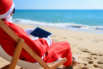 Santa Claus using tablet pc on beach tropical background. Modern internet technology holidays. Back side view of person doing xmas education project, take self photo, relaxing, play gadget or shopping