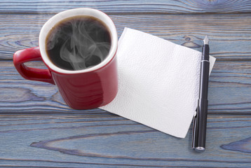 Red cup of hot coffee with a napkin on the table. A pen. Place for text