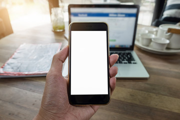 Close up of a man holding smartphone with blank screen mobile and laptop computer at coffee shop.