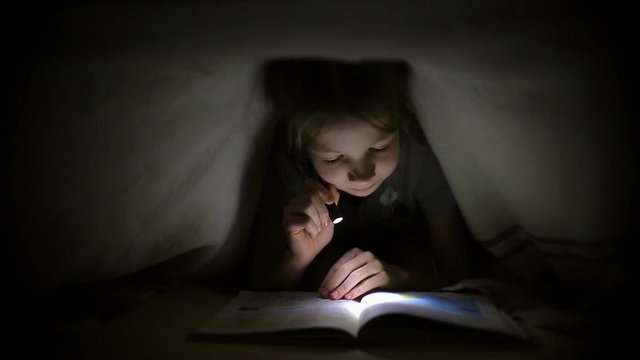 little girl is reading a book under a blanket with a flashlight in a dark room at night