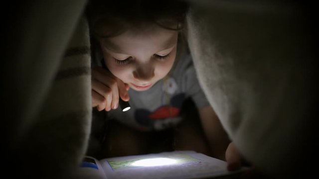 little girl is reading a book under a blanket with a flashlight in a dark room at night