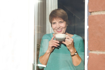 Senior woman looking at the window drinking a warm beverage, coffee or tea