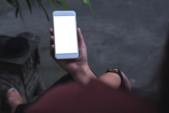 Mockup image of a woman sitting crossed leg and holding white mobile phone with blank screen with concrete floor background