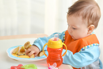 Cute little baby eating sliced fruits at table in room