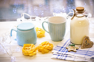 A beautiful New Year's still-life. On the table is a cup of cappuccino and a bottle of milk. Christmas decoration of cookies in the form of a heart and a star. In the background, glowing garlands.