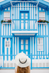 Fototapeta premium Young woman in sun hat standing with old traditional house facade with blue stripes on the background in Costa Nova resort in Portugal