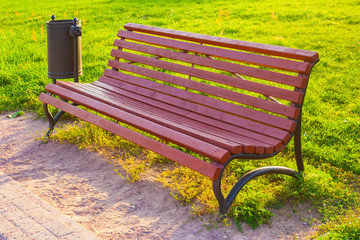 Wooden bench with wrought-iron legs and metal black trash can in the park on a background of green lawn