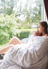 young smiling woman sits on a windowsill with a cup of coffee. the woman is sitting without clothes, wrapped in a blanket