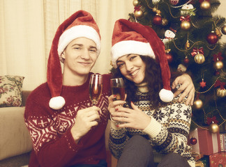Happy couple sit near christmas tree with glass of champagne. Winter holiday and love concept. Yellow toned