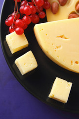 Hard cheese with holes on a black plate, cheese with red berries, peanuts, viburnum, cheese in minimalism style, cheese and red berries on a dark blue background