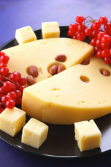 Hard cheese with holes on a black plate, cheese with red berries, peanuts, viburnum, cheese in minimalism style, cheese and red berries on a dark blue background