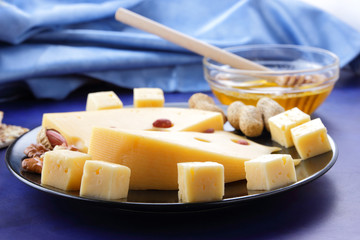 Hard cheese with holes on a black plate, cheese with peanuts, honey, biscuits, walnuts, cheese in minimalism style, cheese on dark blue background in retro style