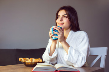 Beautiful caucasian girl in a white bathrobe enjoys morning coffee and croissant, time in the kitchen