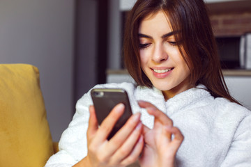 Cheerful woman wears white bathrobe sitting in yellow armchair on the morning kitchen holding smartphone