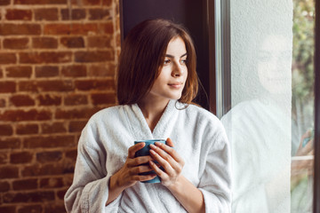 Beautiful caucasian girl, in a white bathrobe, enjoys morning coffee time in the kitchen near window