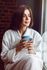 Beautiful caucasian girl, in a white bathrobe, enjoys morning coffee time in the kitchen near window