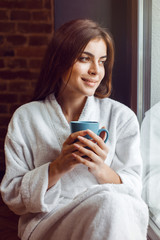 Beautiful caucasian girl, in a white bathrobe, enjoys morning coffee time in the kitchen near window