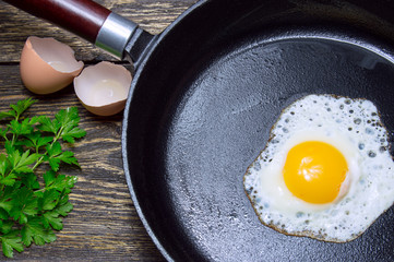 Fried egg in a cast iron pan with parsley on wooden background with eggshell