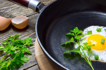 Fried egg with leaves of green onions in a cast iron pan with parsley on wooden background with eggshell