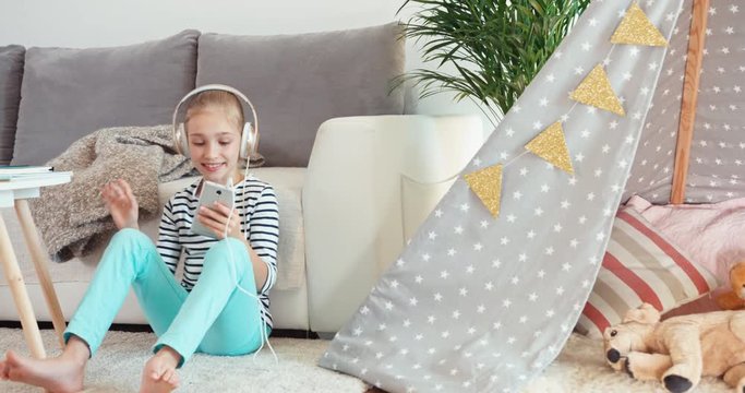 Child girl listening music from headphones from cell phone sitting on the white carpet near sofa in the living room