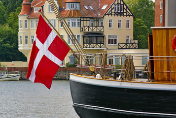 Stern of a traditional sailing ship with large danish flag hanging from a flagpole and waterfront buildings in the background
