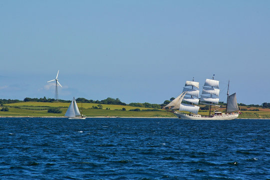 Traditional square-rigged tall ship and modern two-masted yacht at sea in front of a green seashore with fields and wind power station