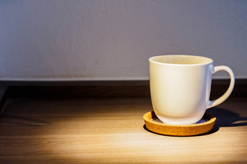 A cup of coffee is placed on a wooden floor under the light of a lamp.